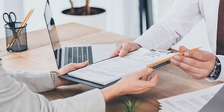 Persoon geeft papier en potlood aan iemand aan de overkant, met een bureaublad en laptop op de achtergrond
