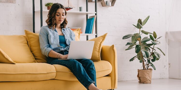 Vrouw zittend op gele bank met laptop op school. Naast de bank staat een plant