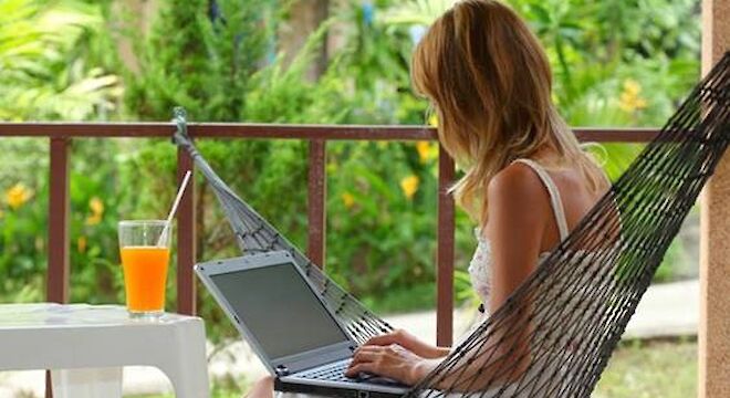 Vrouw zit in hangmat en tikt op laptop. Op de voorgrond staat een tafel met een cocktail daarop.