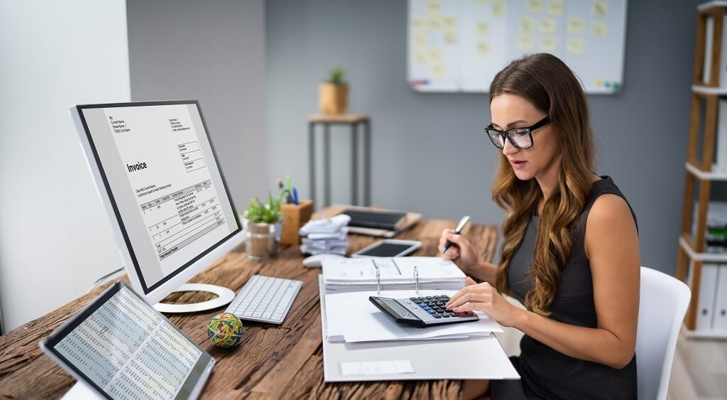 vrouw achter bureau met laptop, tablet en een map met een rekenmachine waar ze iets op uitrekent