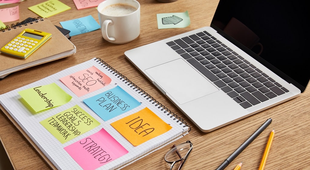 Bureaublad met laptop en een notitieboekje met post-its erop geplakt.