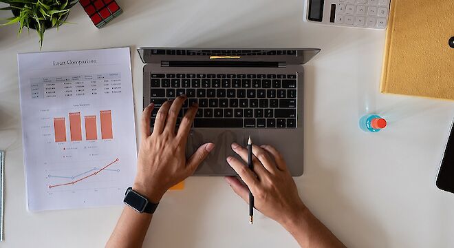 Bovenaanzicht 2 handen die tikken op laptop. Een hand met pen tussen vingers. Op bureaublad ook papier met grafieken erop.