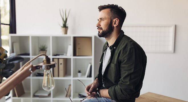 Man zittend op bureaublad met bril in hand kijkend naar buiten. Op achtergrond een vakkenkast en op voorgrond een lamp