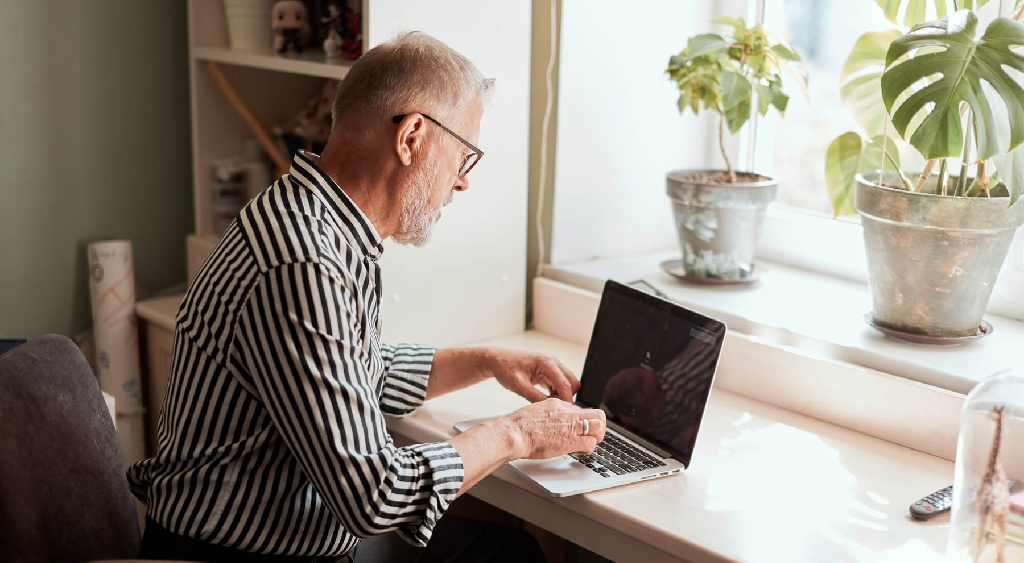 Man met bril zittend aan bureau met laptop voor hem. Op vensterbank staan planten.