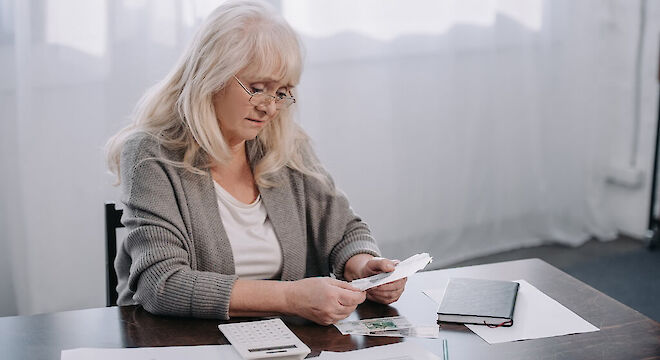 Oudere vrouw aan bureau die kijkt naar briefjes in hand.