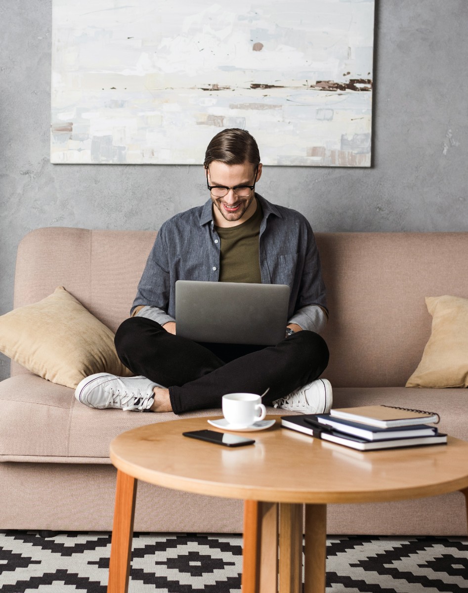 Man zit in kleermakerszit op de bank met laptop in hand, voor hem staat een houten tafel met notitieboekjes, een kop koffie en een telefoon.